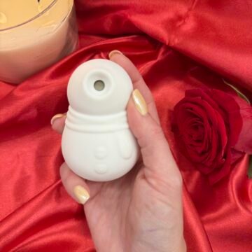 Snowmen suction vibrator in while silicone, held in hand on red satin background with rose and candle